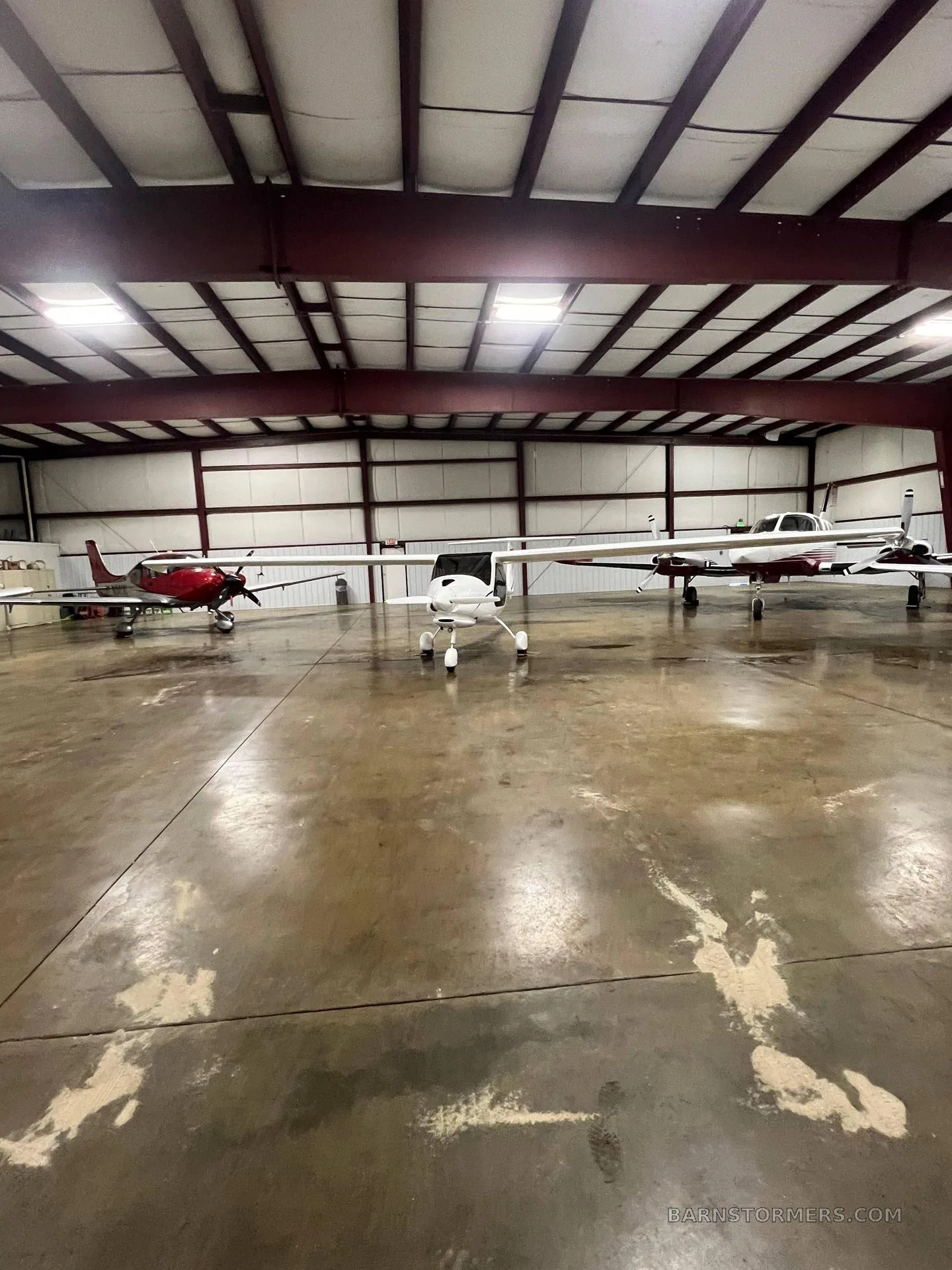 Three light aircraft, including a white Pipistrel Sinus, are parked inside a large hangar with a concrete floor.