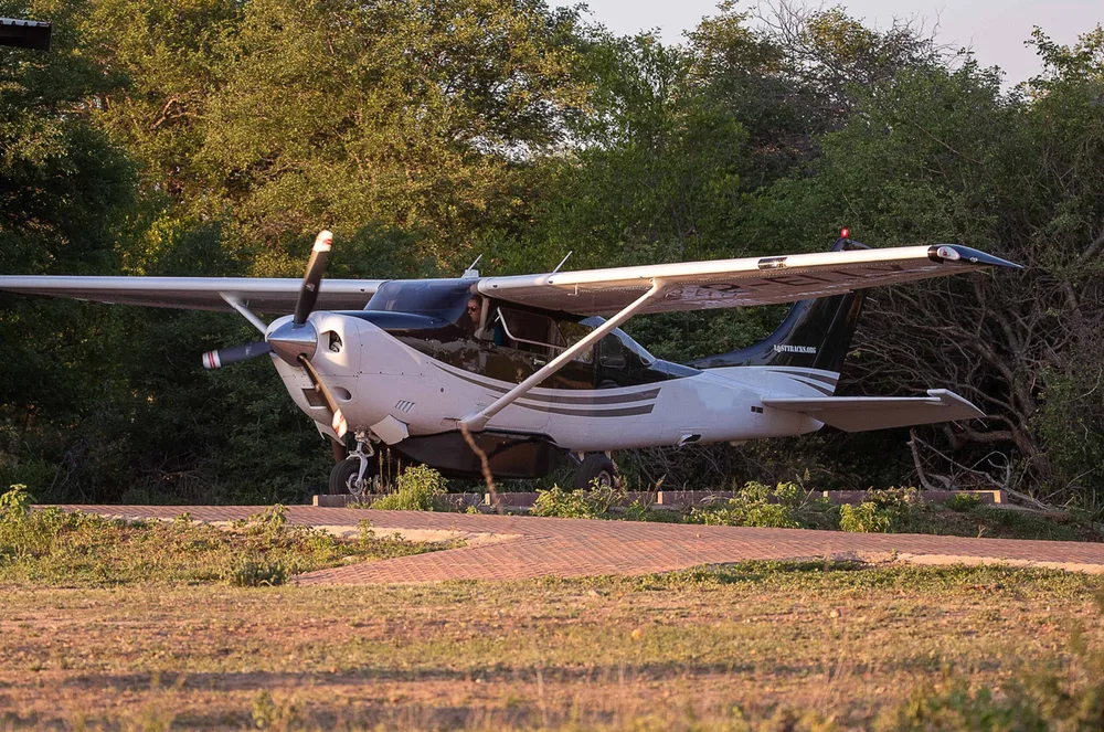 A black and white single-engine propeller aircraft is seen from the front-left, on a paved path surrounded by green trees.