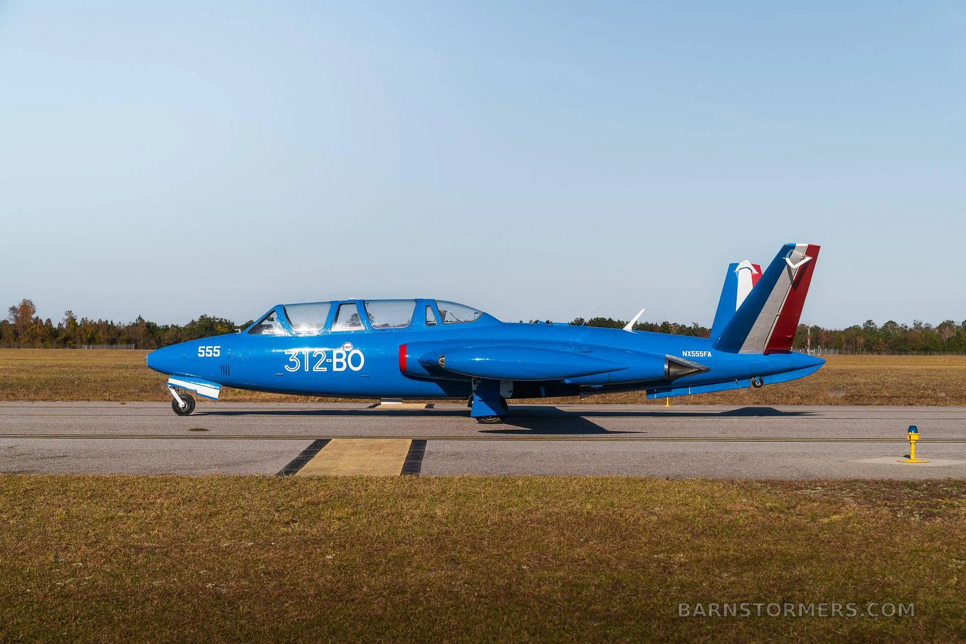 A blue Fouga CM.170 Magister jet aircraft with closed canopies is shown from the side on an asphalt runway.