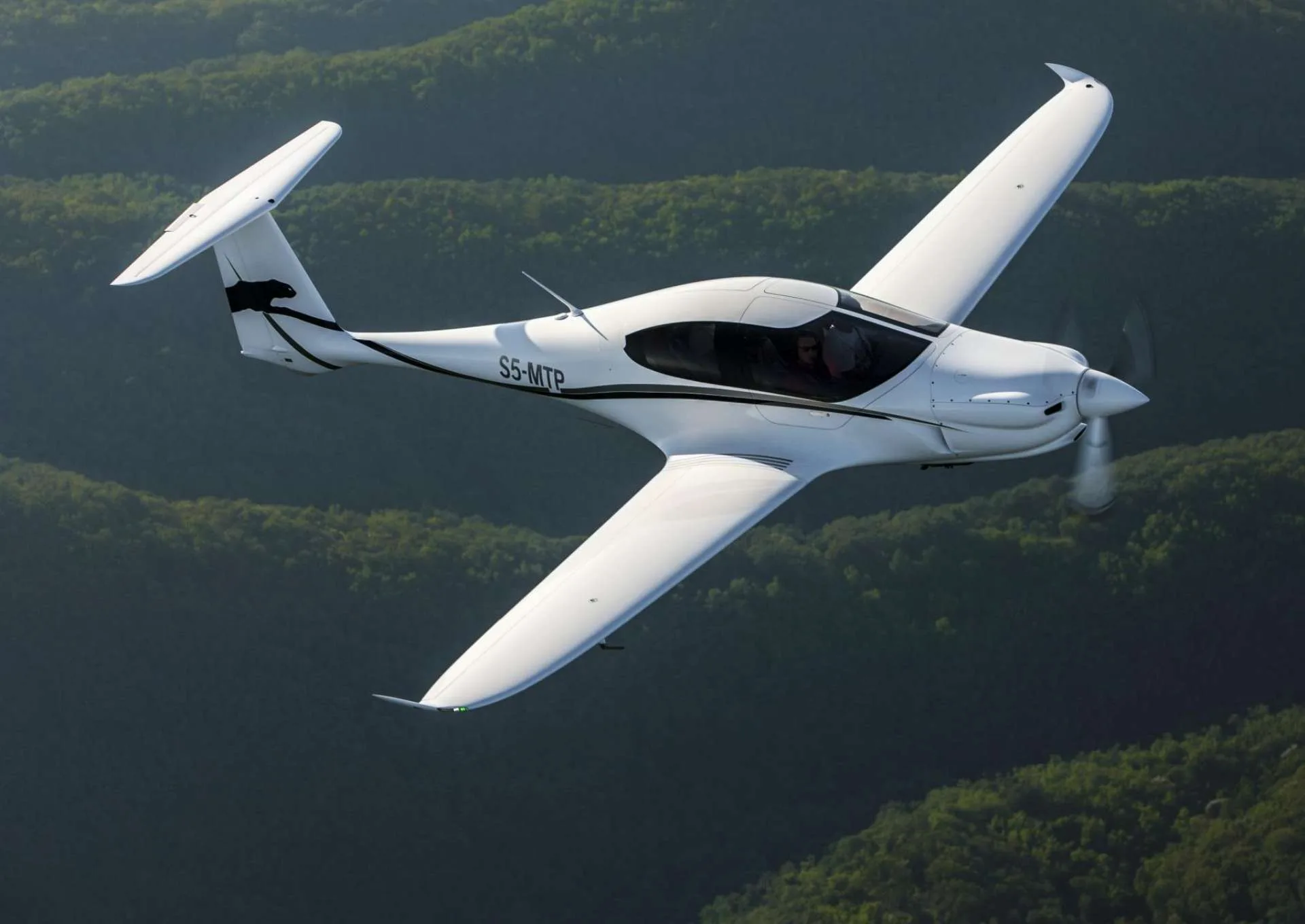 A white single-engine propeller aircraft with black accents and a panther logo on the tail flies over green forested hills.