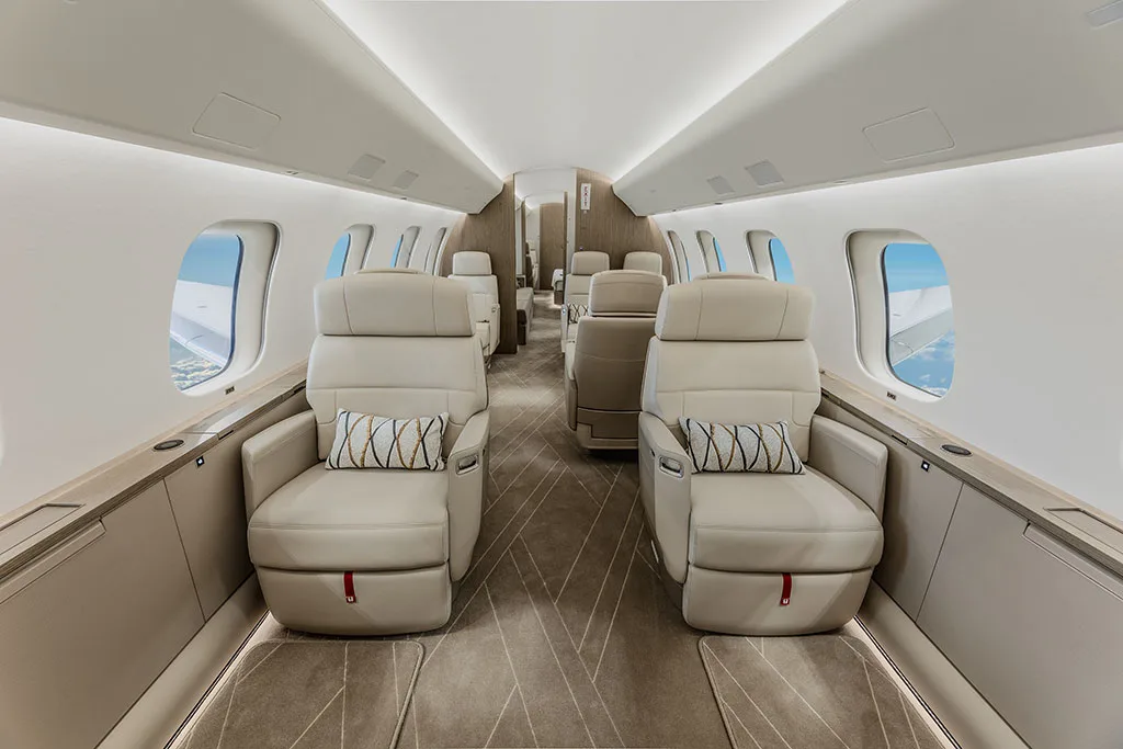 A view inside a private jet cabin showing two rows of beige leather seats with decorative pillows.