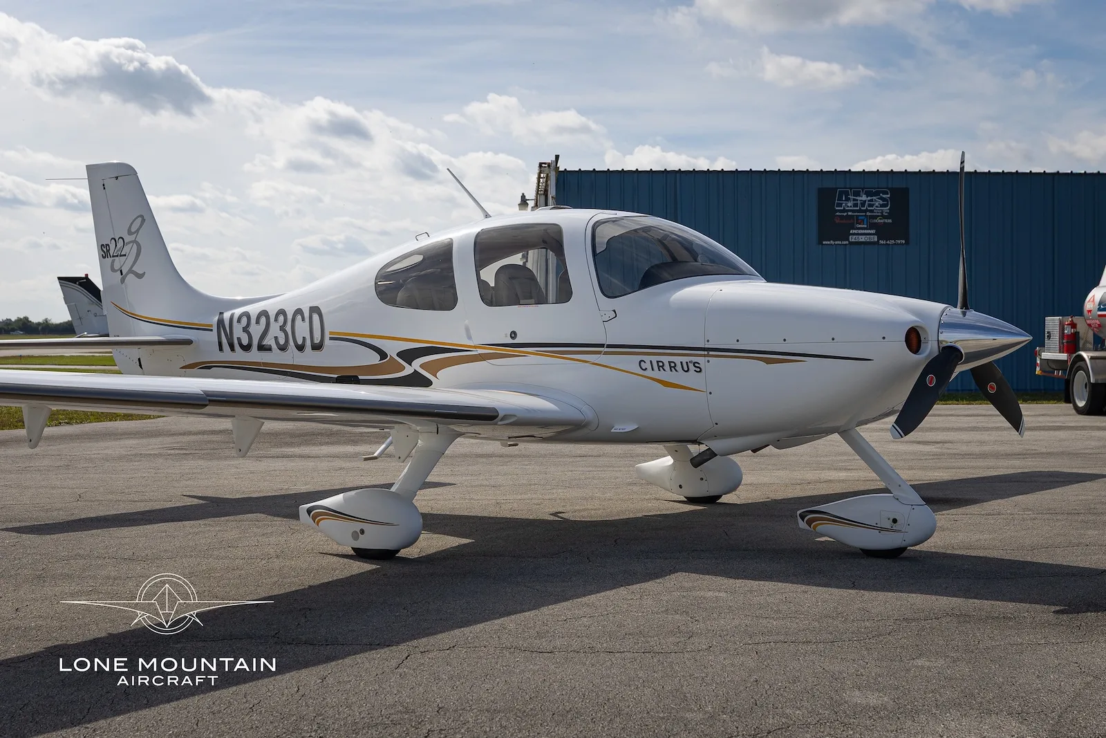 A white Cirrus SR22 aircraft with gold and black accents, registration N323CD, parked on an airfield with its doors closed.