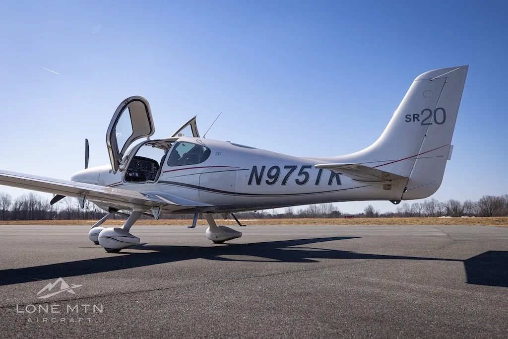 Side view of a silver Cirrus SR20 aircraft with registration N975TK parked on an airfield with its doors open.