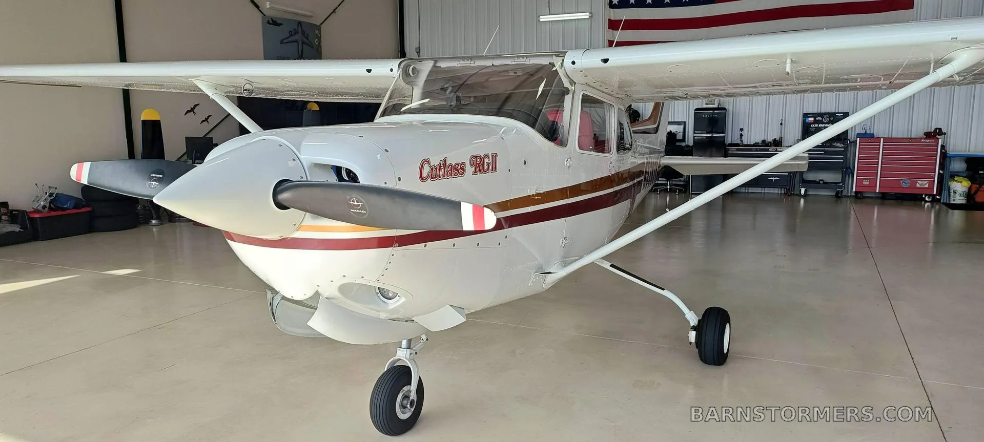 A white Cessna Cutlass RGII (N5393V) with red and gold stripes is shown from a head-on view inside a hangar.