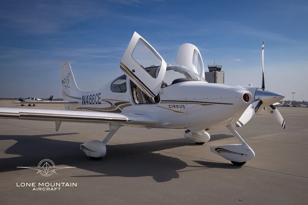 A white Cirrus SR22 aircraft with gold and black stripes, registration N466CE, parked on an airfield with its doors open, viewed from the front-right.