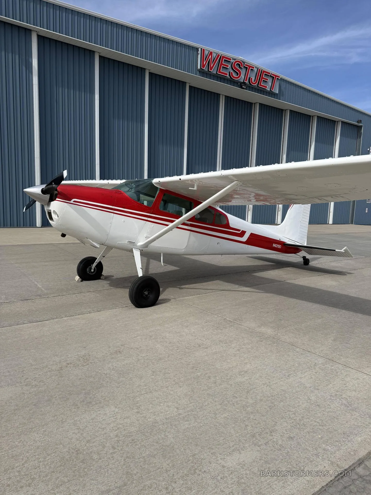 A white and red single-engine propeller aircraft (N42100) parked on concrete in front of a blue Westjet hangar.