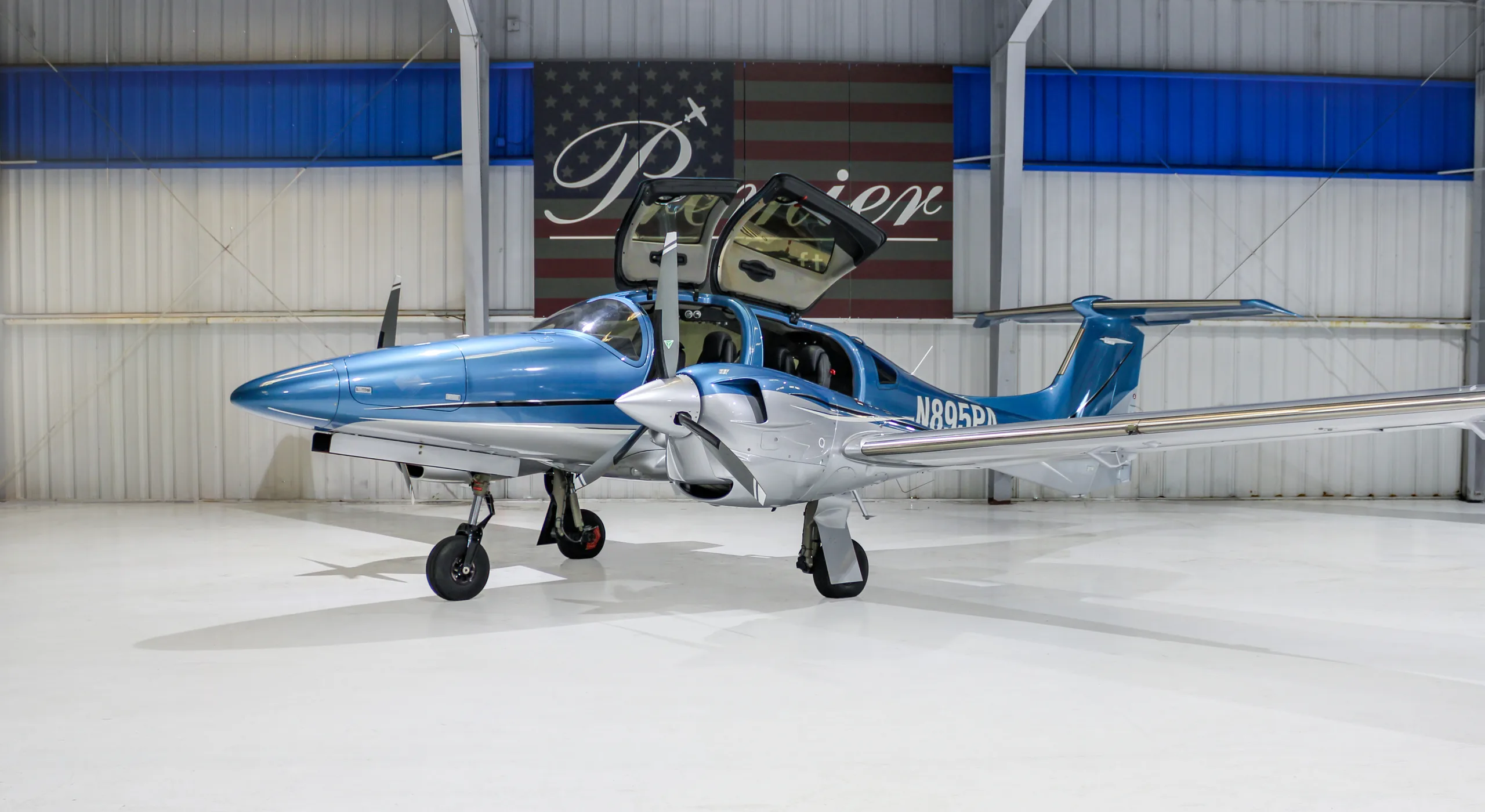 A blue and silver Diamond DA62 aircraft with its doors open, parked in a hangar.