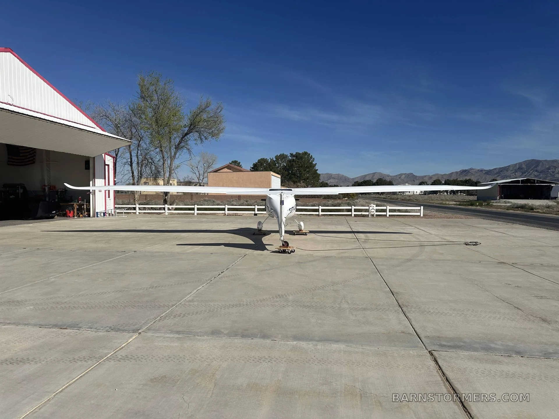 A white Pipistrel Sinus aircraft with blue accents is viewed from the front, parked on an apron in front of a hangar.