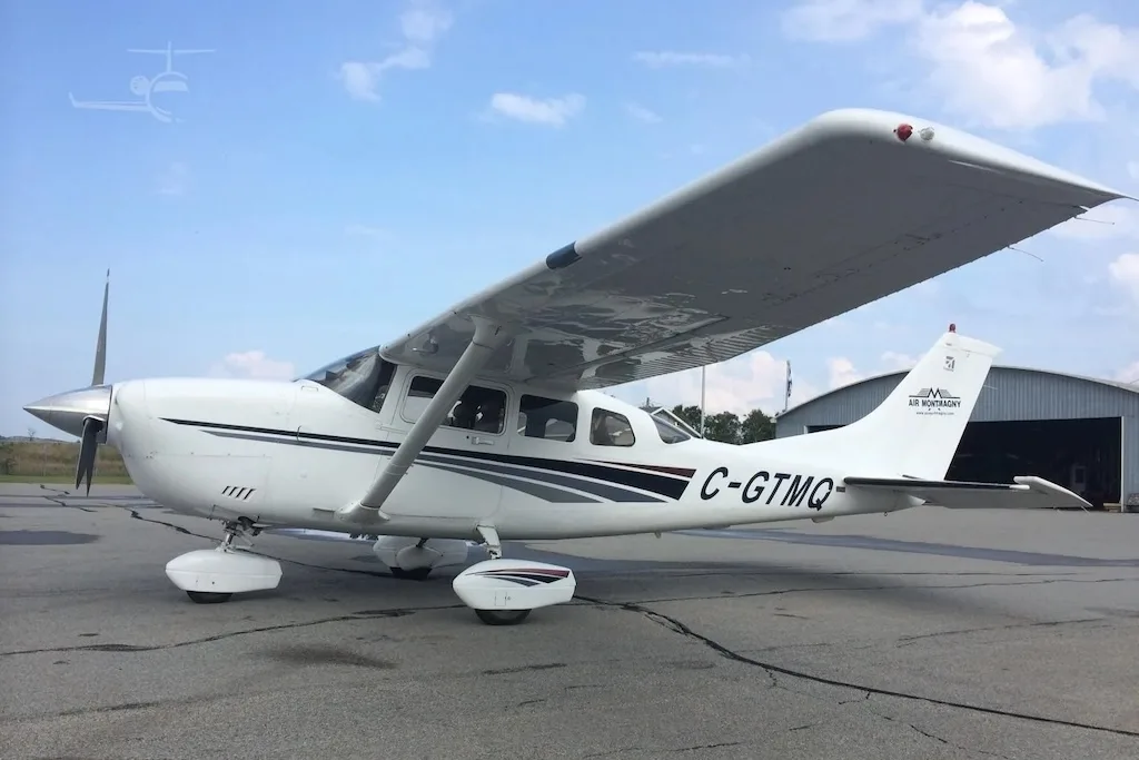 A white single-engine propeller aircraft with black and grey stripes, registration C-GTMQ, parked on an asphalt tarmac under a partly cloudy sky.