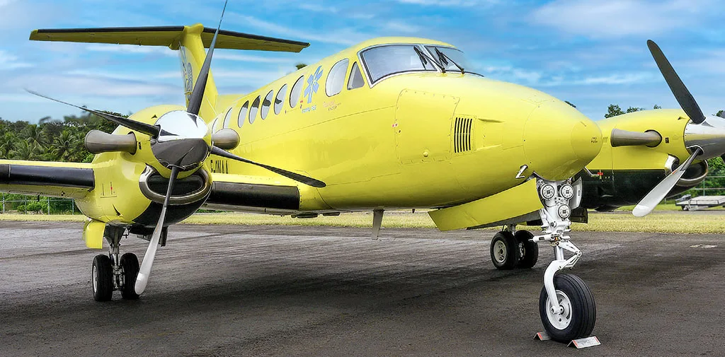 A bright yellow twin-engine turboprop aircraft with an ambulance logo on the side, parked on an asphalt runway under a blue sky.