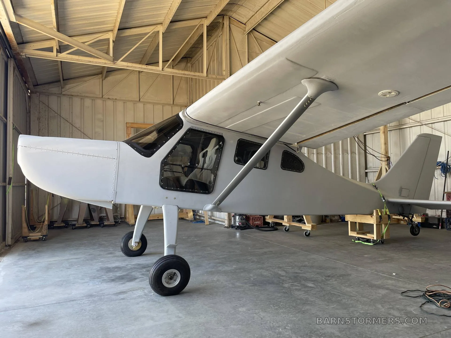 A white, high-wing aircraft with a taildragger configuration is parked inside a hangar.