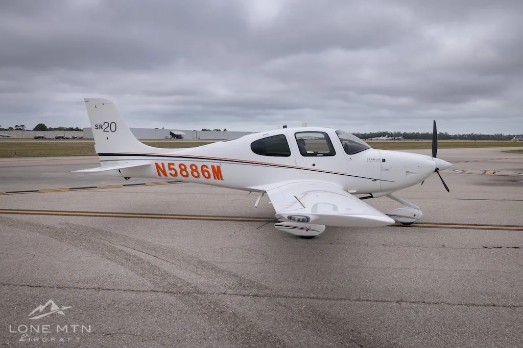 A three-quarter front view of a white Cirrus SR20 aircraft with orange and black stripes, registration N5886M, on an airfield.