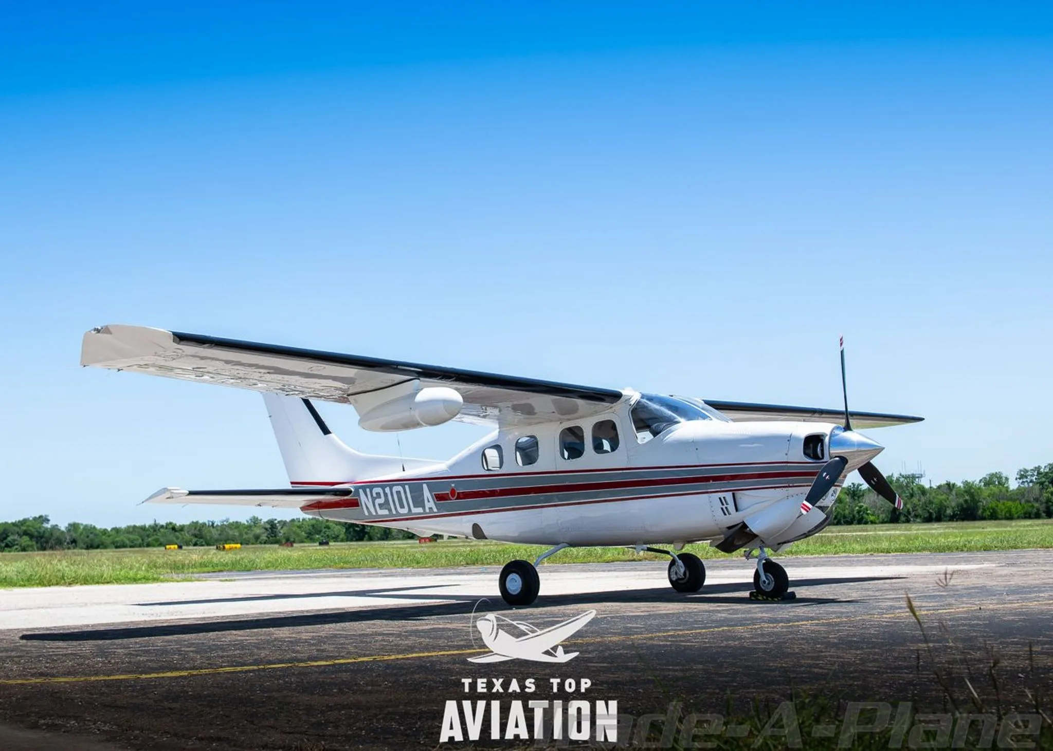 A white Cessna 210 Centurion (N210LA) with red and gray stripes parked on an airfield under a clear blue sky.