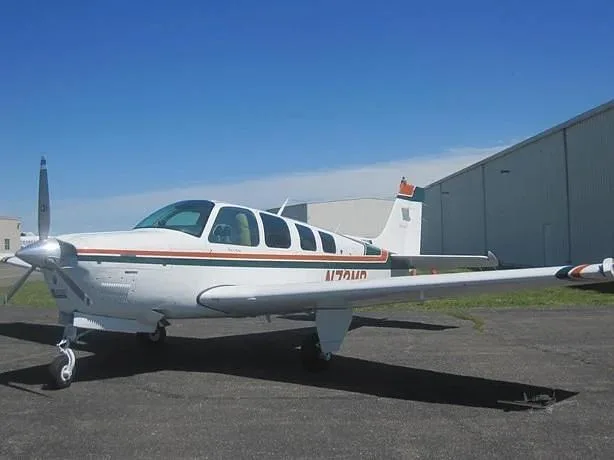 A white Beechcraft Bonanza N72MD with orange and green stripes is parked on an airfield tarmac under a clear blue sky.