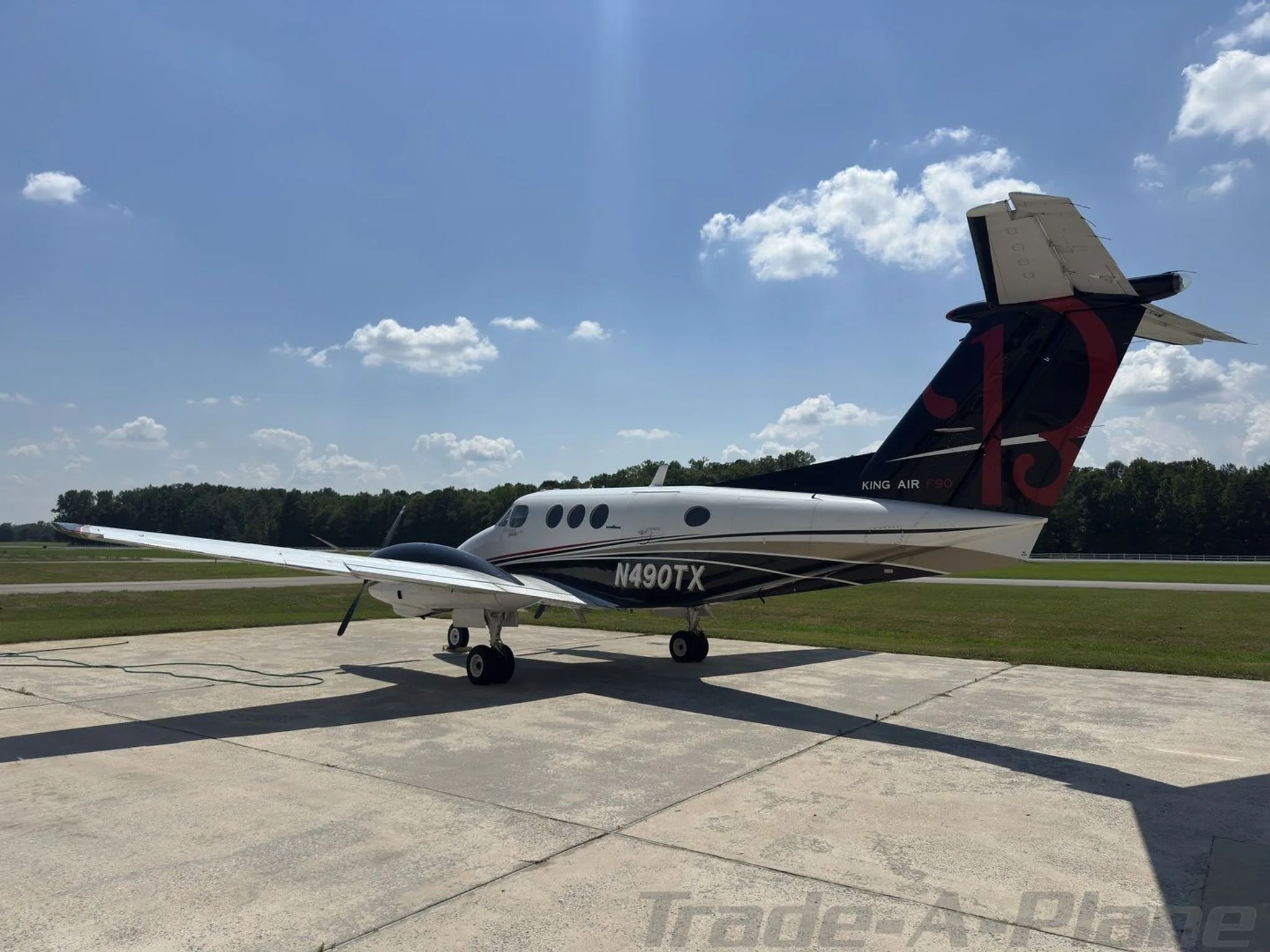 A rear three-quarter view of a white and black Beechcraft King Air F90, N490TX, parked on a tarmac with grass and trees in the background.