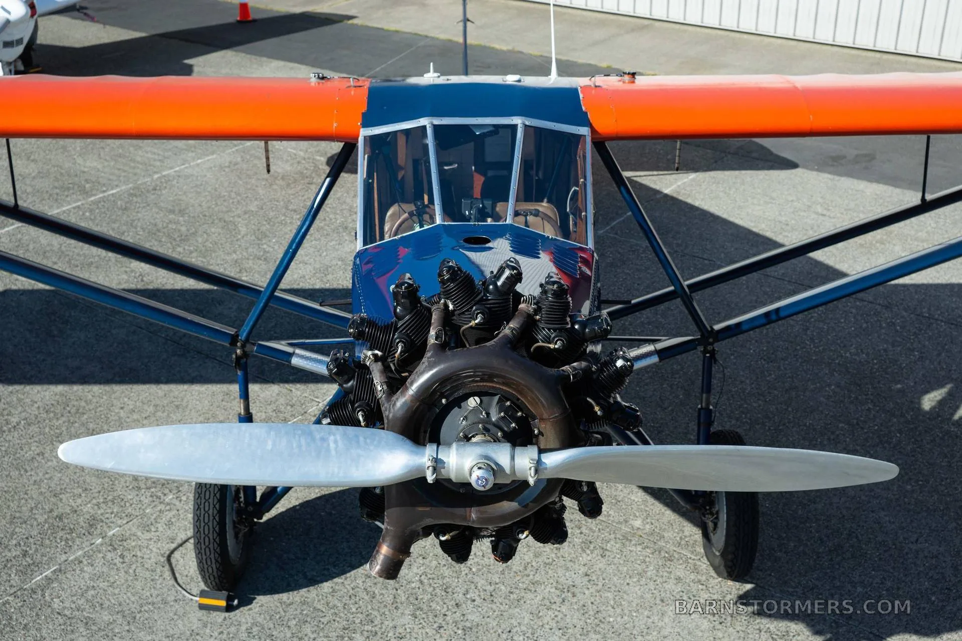 A close-up front view of a vintage blue and orange aircraft, highlighting its radial engine and propeller.