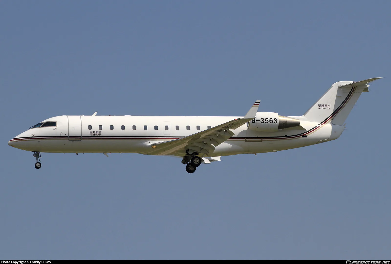 A white Astro Air Bombardier Challenger 850 aircraft with registration B-3563 is captured in flight against a clear blue sky, with its landing gear extended.