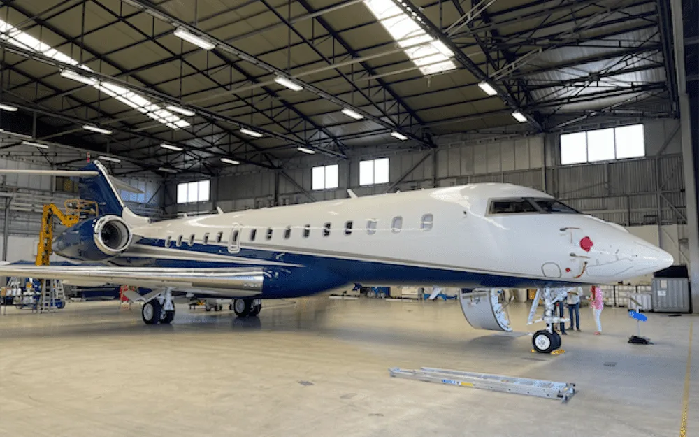 A white and blue business jet, likely a Bombardier Global, parked inside a hangar.