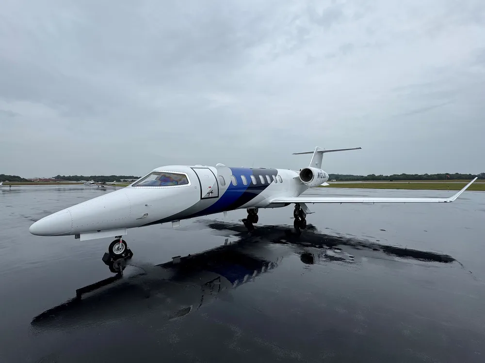 A white business jet with blue and grey accents, registration N12JS, parked on a wet tarmac under a cloudy sky.
