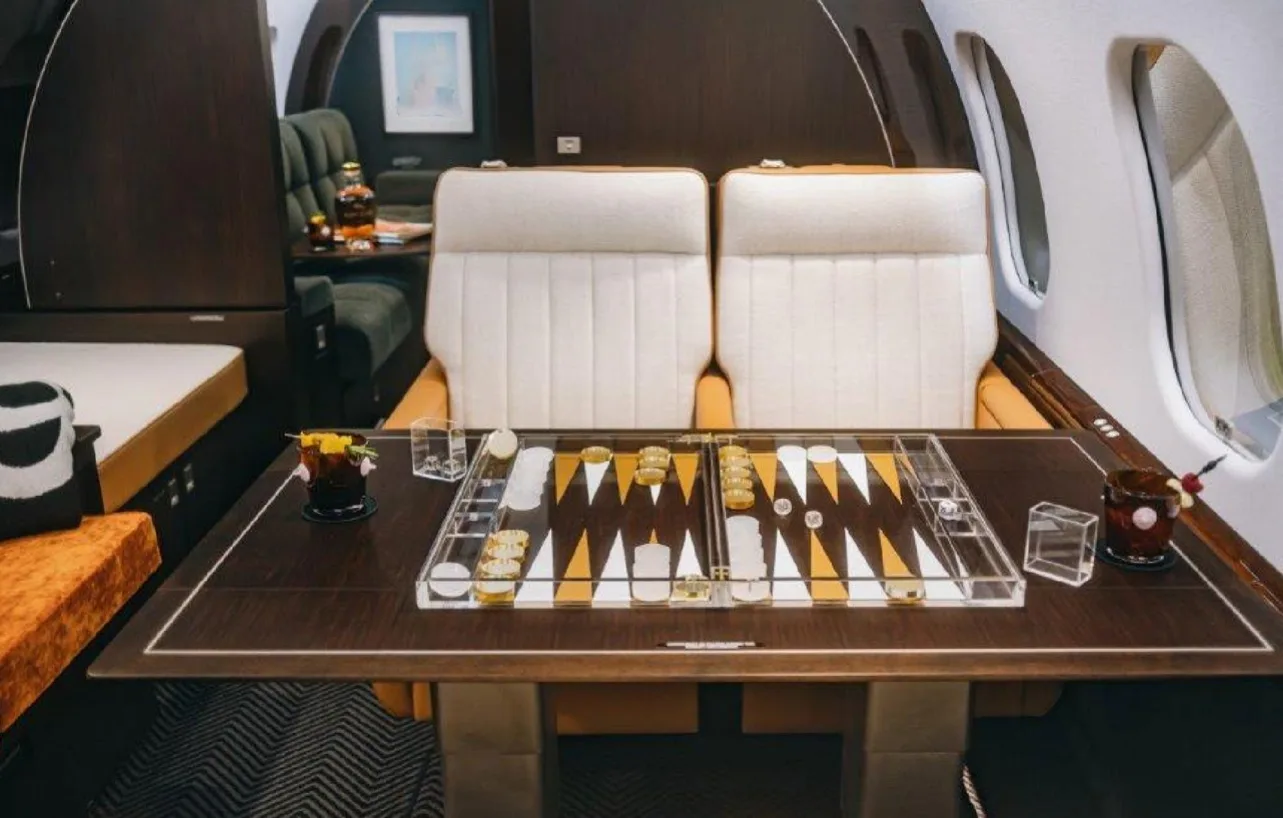 The luxurious interior cabin of a private jet, featuring two cream and tan seats facing a table with a backgammon set.