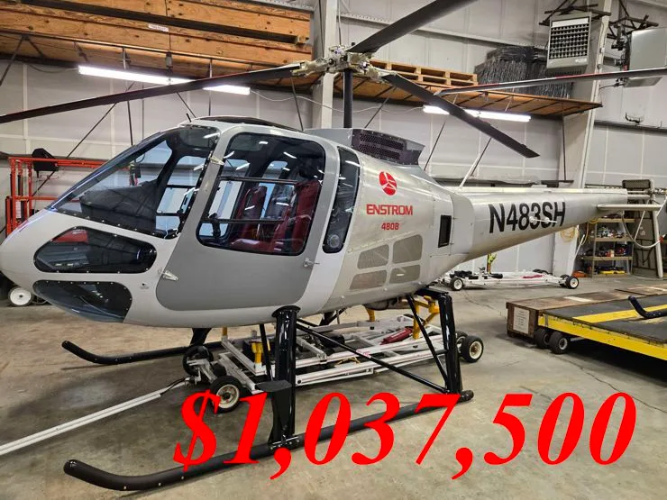 A light grey Enstrom 480B helicopter with red interior seats, parked inside a hangar.