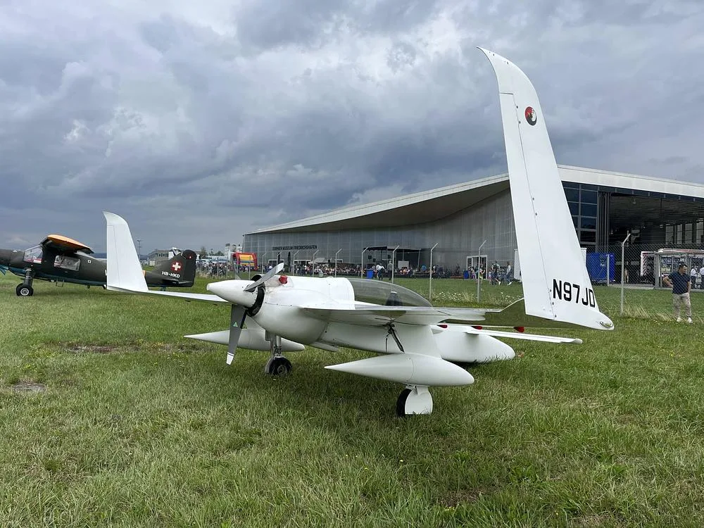A white, single-engine aircraft with registration N97JD is parked on a grassy field next to a large modern building and other aircraft.