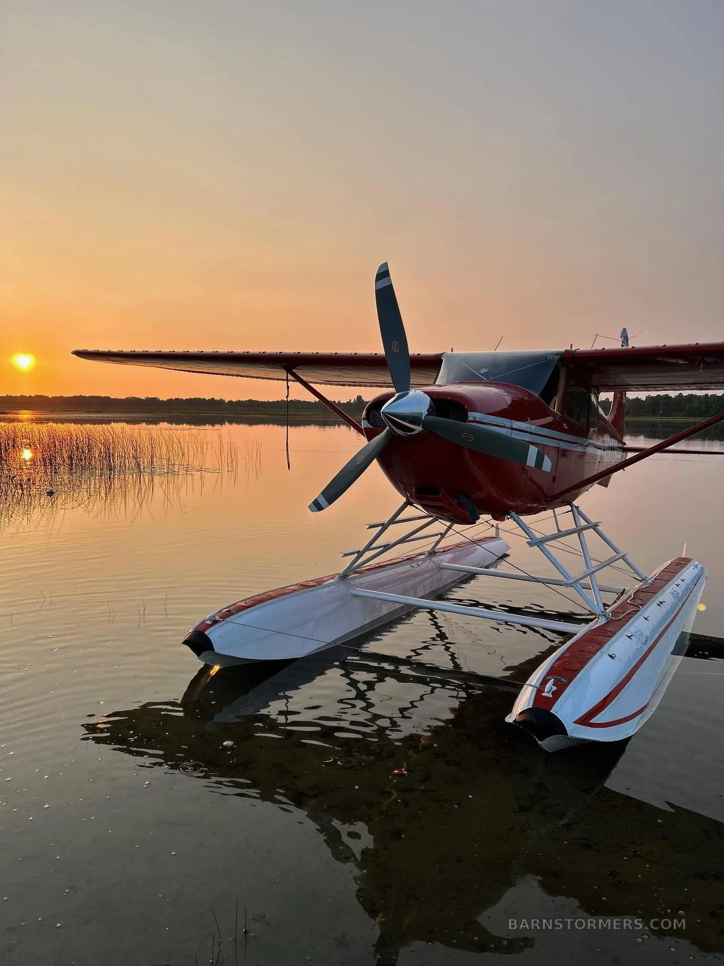 A red floatplane with white floats resting on the water at sunset.