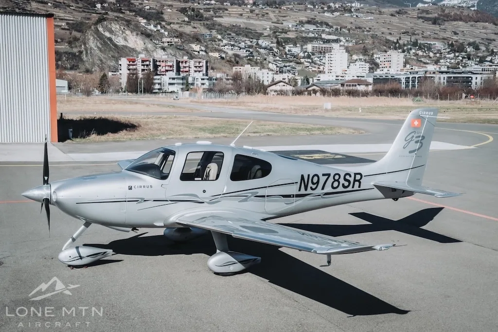 A silver Cirrus SR22 aircraft (N978SR) parked on an airfield with a town and hills in the background.