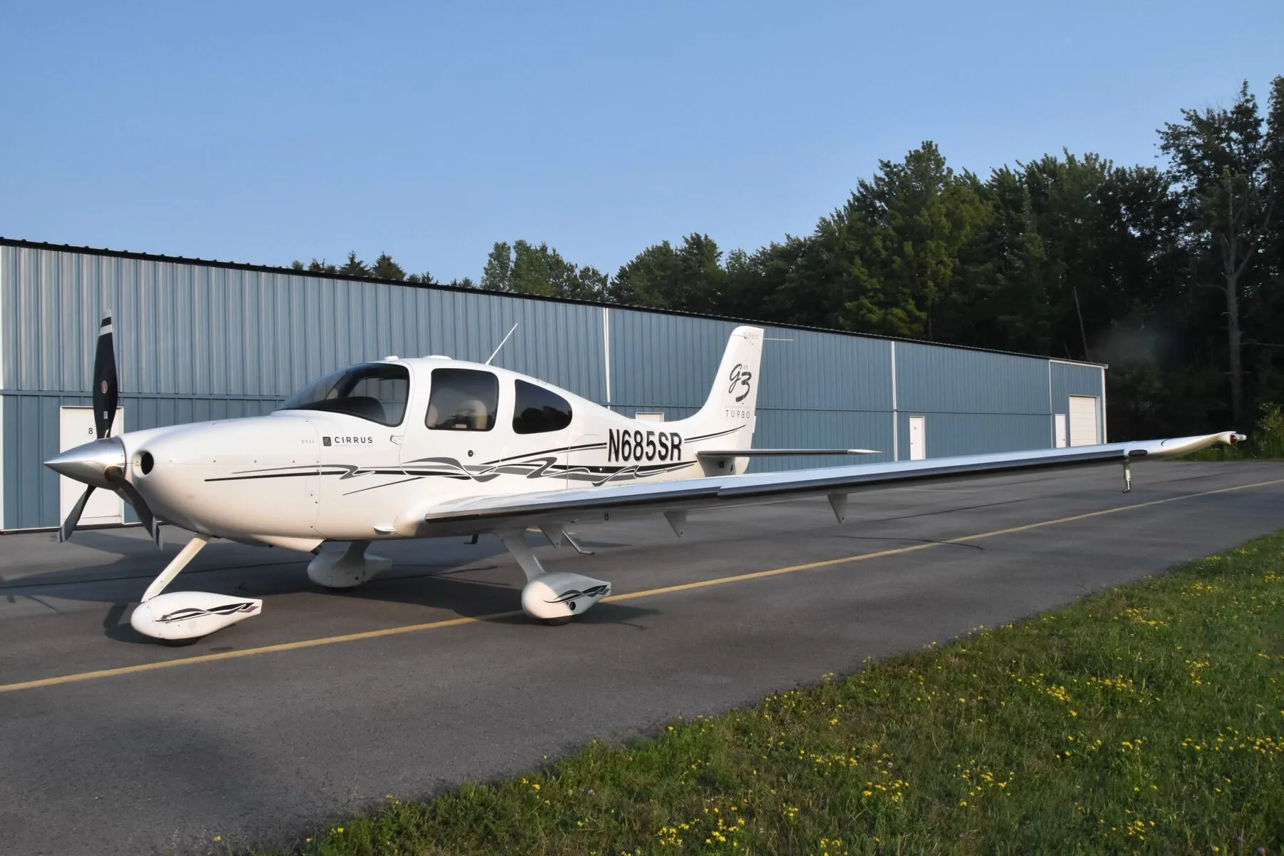A white Cirrus SR22T aircraft (N685SR) parked on an airfield with a hangar and trees in the background.