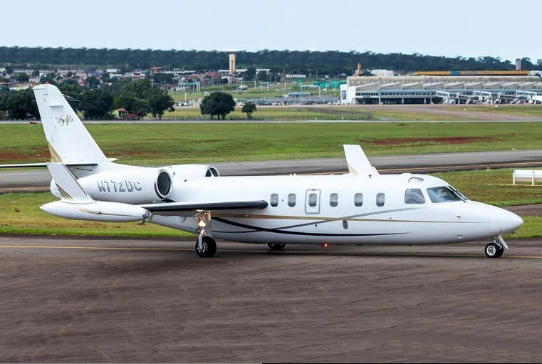 A white business jet with gold and grey stripes, registration N772DC, on an airport tarmac with buildings in the background.