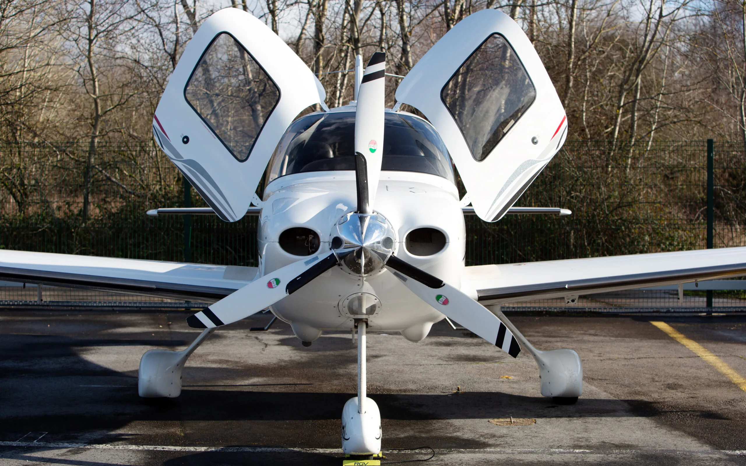 A white Cirrus SR22 G3 aircraft with red and black stripes, registration G-FELC, parked head-on on a tarmac with its gull-wing doors open.