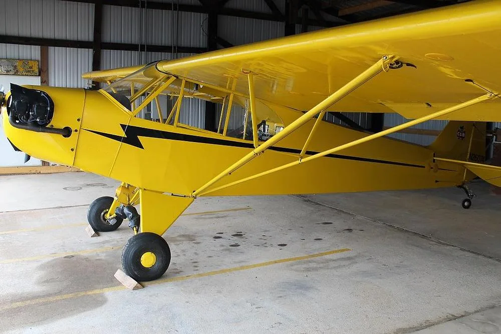 A yellow Piper Cub-style aircraft with a black lightning bolt stripe is parked in a hangar, viewed from the front-left side.