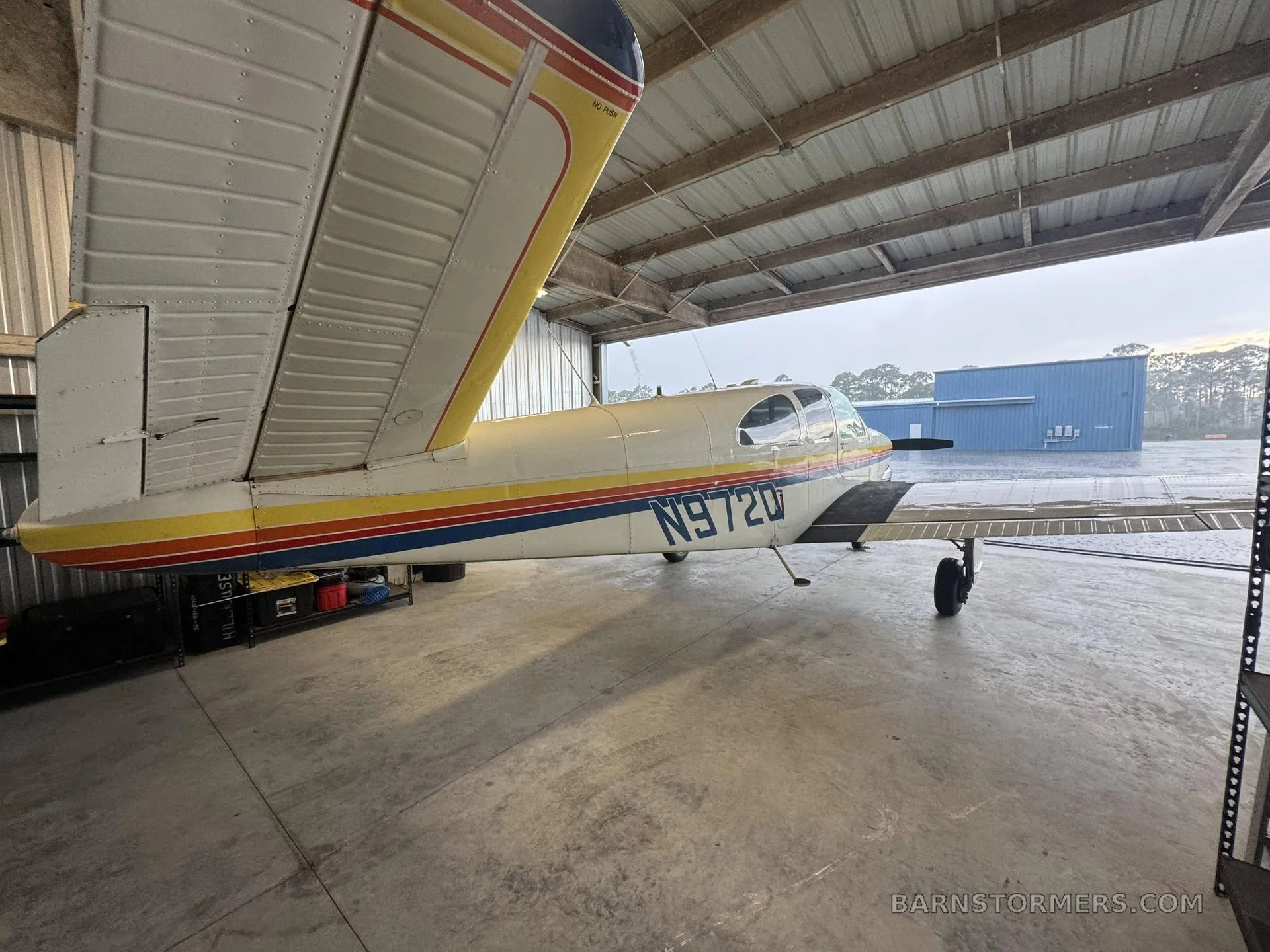 A side view of a white aircraft with rainbow stripes and yellow wings, parked inside a hangar with a view of a blue building outside.