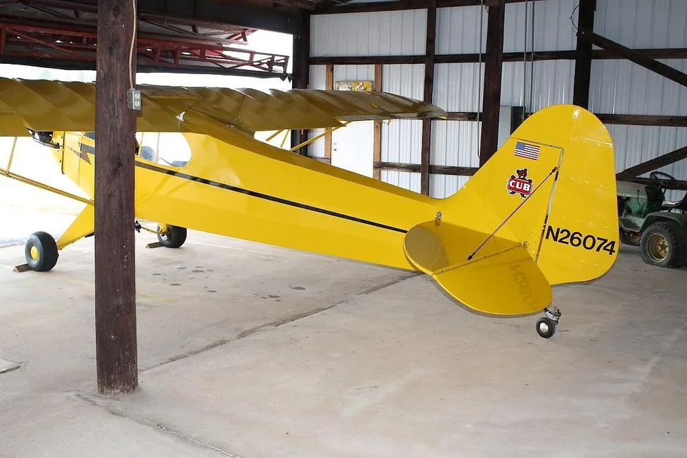 The rear-right view of a yellow Piper Cub-style aircraft with registration N26074 parked in a hangar.