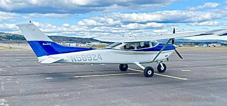 A white and blue Cessna 182 Skylane (N58924) parked on an airfield with mountains in the background.