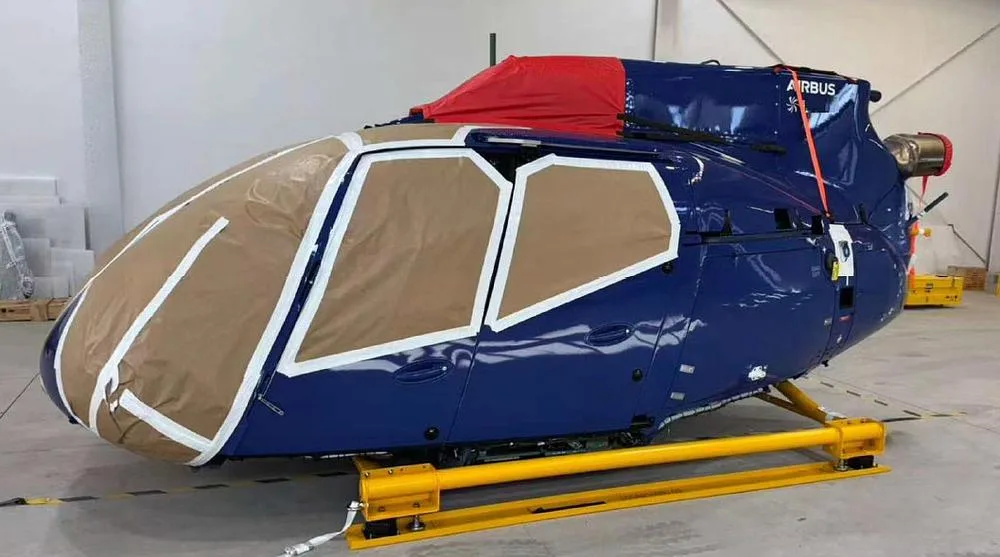The blue fuselage of an Airbus H135 helicopter, with windows and parts covered, on a yellow transport frame in a hangar.