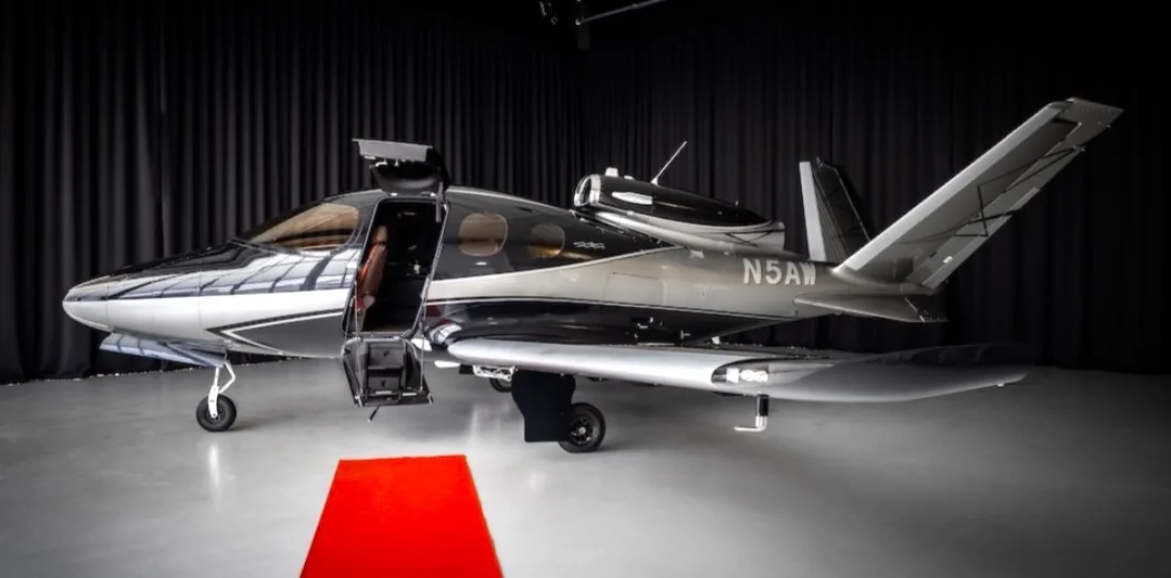A two-tone grey and black Cirrus Vision Jet (N5AW) with its main door open, parked on a grey floor with a red carpet, against a dark curtain background.