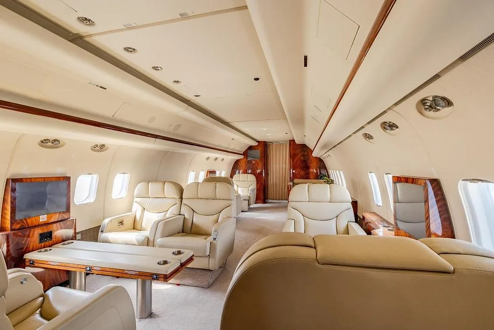 A luxurious aircraft interior with cream leather seats, wood tables, and polished wood paneling.