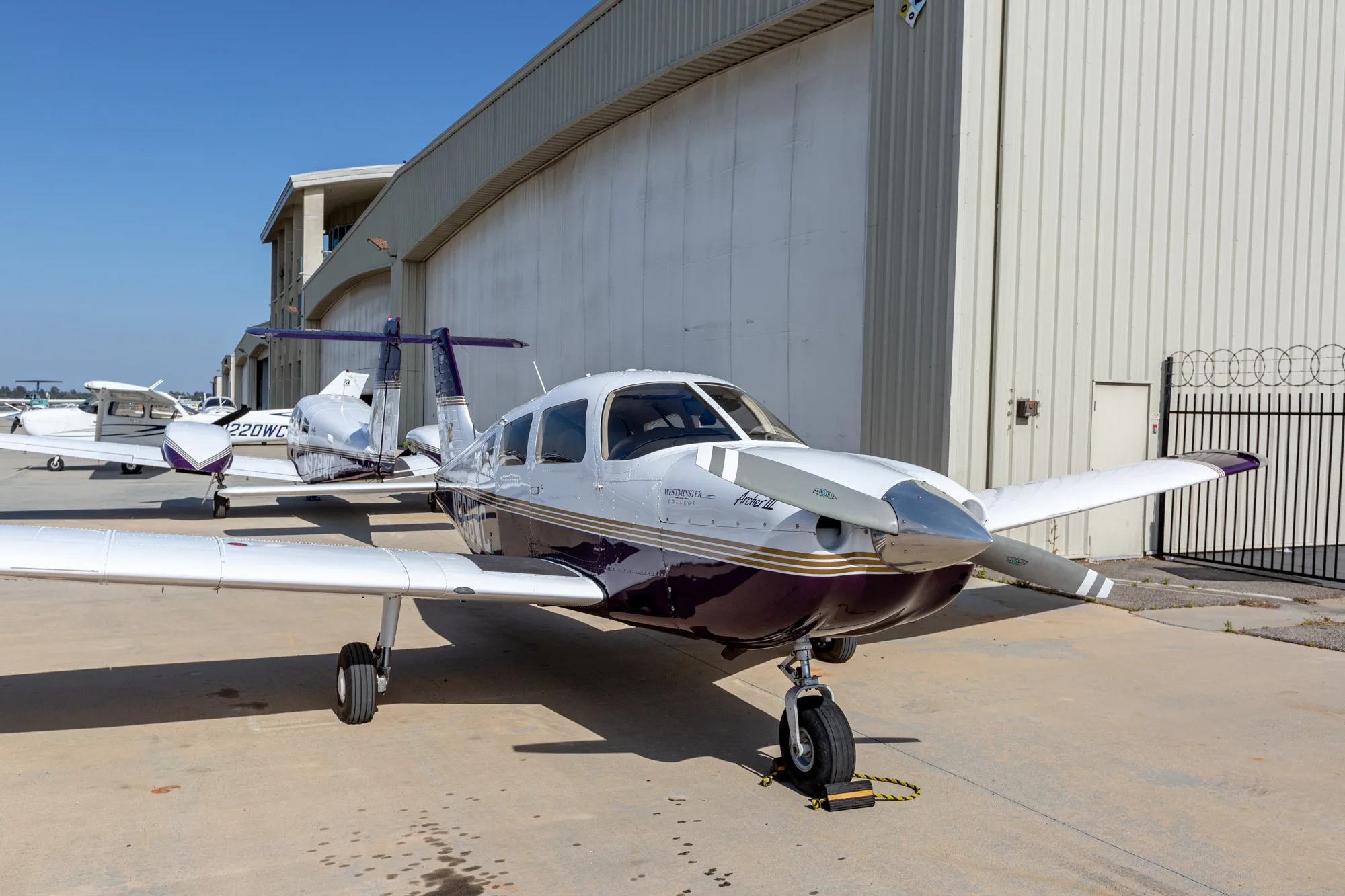 A white and purple Piper Archer III aircraft, N968WC, parked on an airfield in front of hangars.