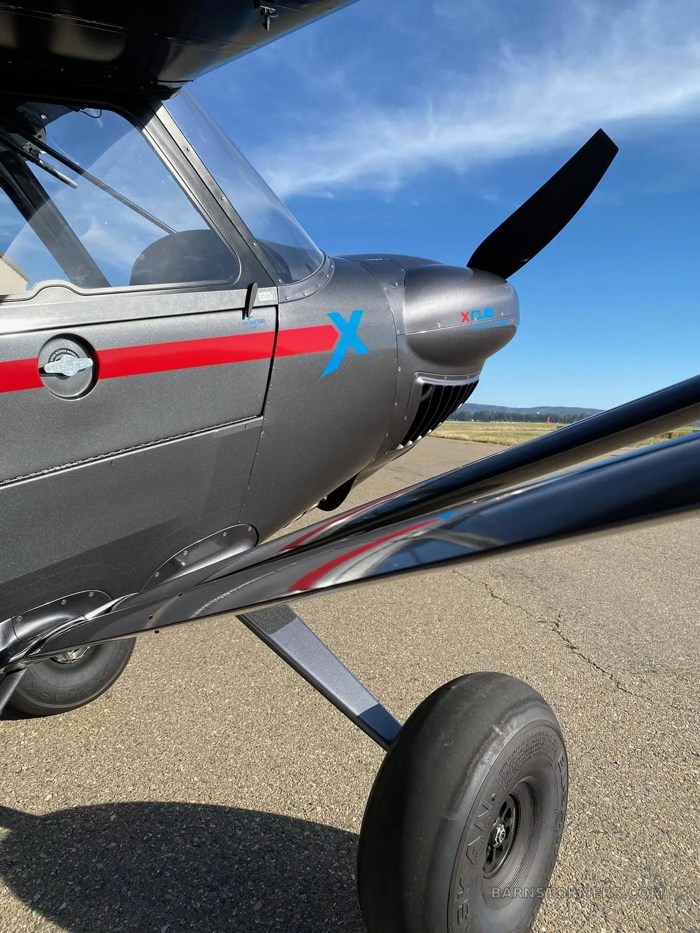 A close-up view of the front fuselage and propeller of a dark gray X-Cub aircraft with a red stripe and blue 'X' logo.