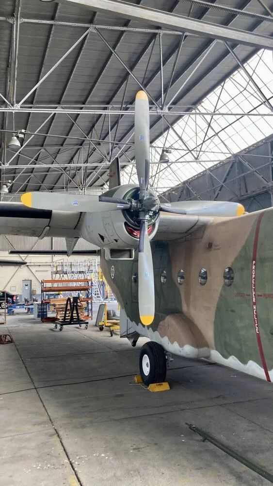 A side view of a camouflage military transport aircraft's propeller and engine in a hangar.