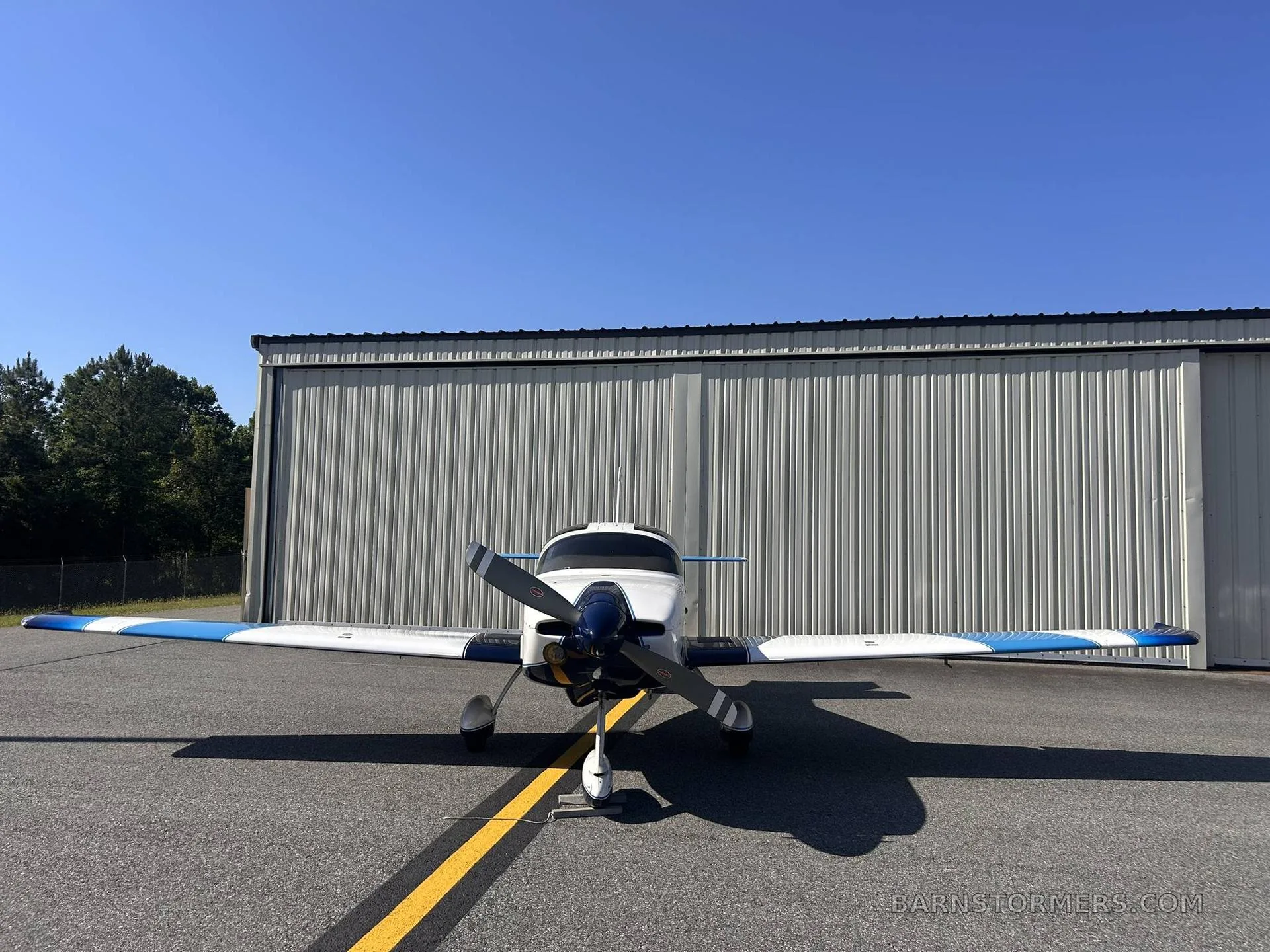 A white and blue single-engine aircraft is parked on an asphalt tarmac in front of a hangar, viewed from the front.