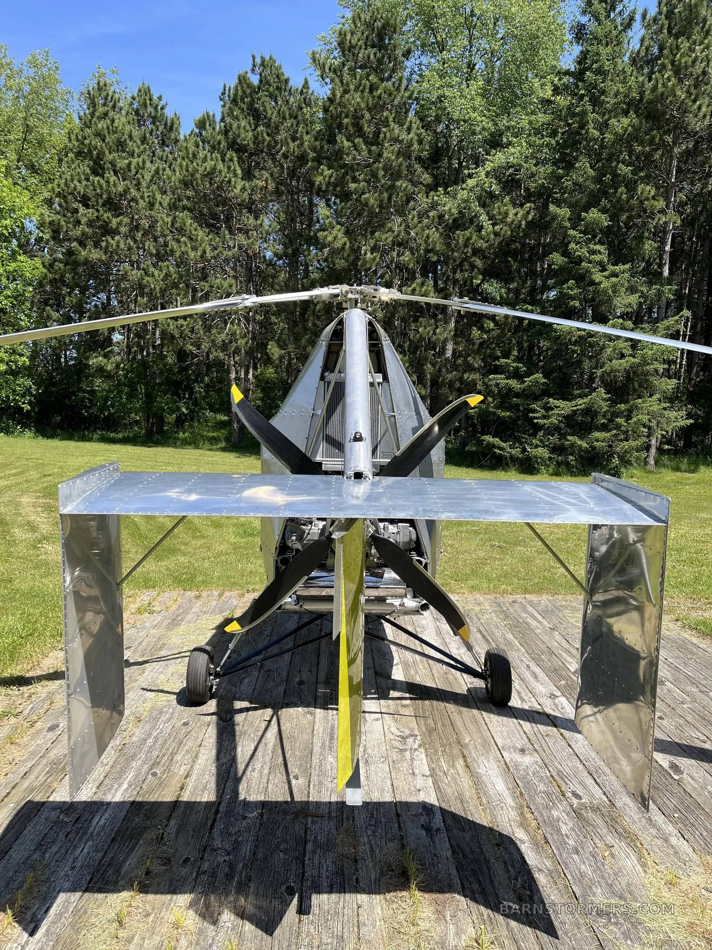 A front view of a silver single-seat ultralight helicopter/autogyro with a yellow propeller, parked on a wooden pad in a grassy field.
