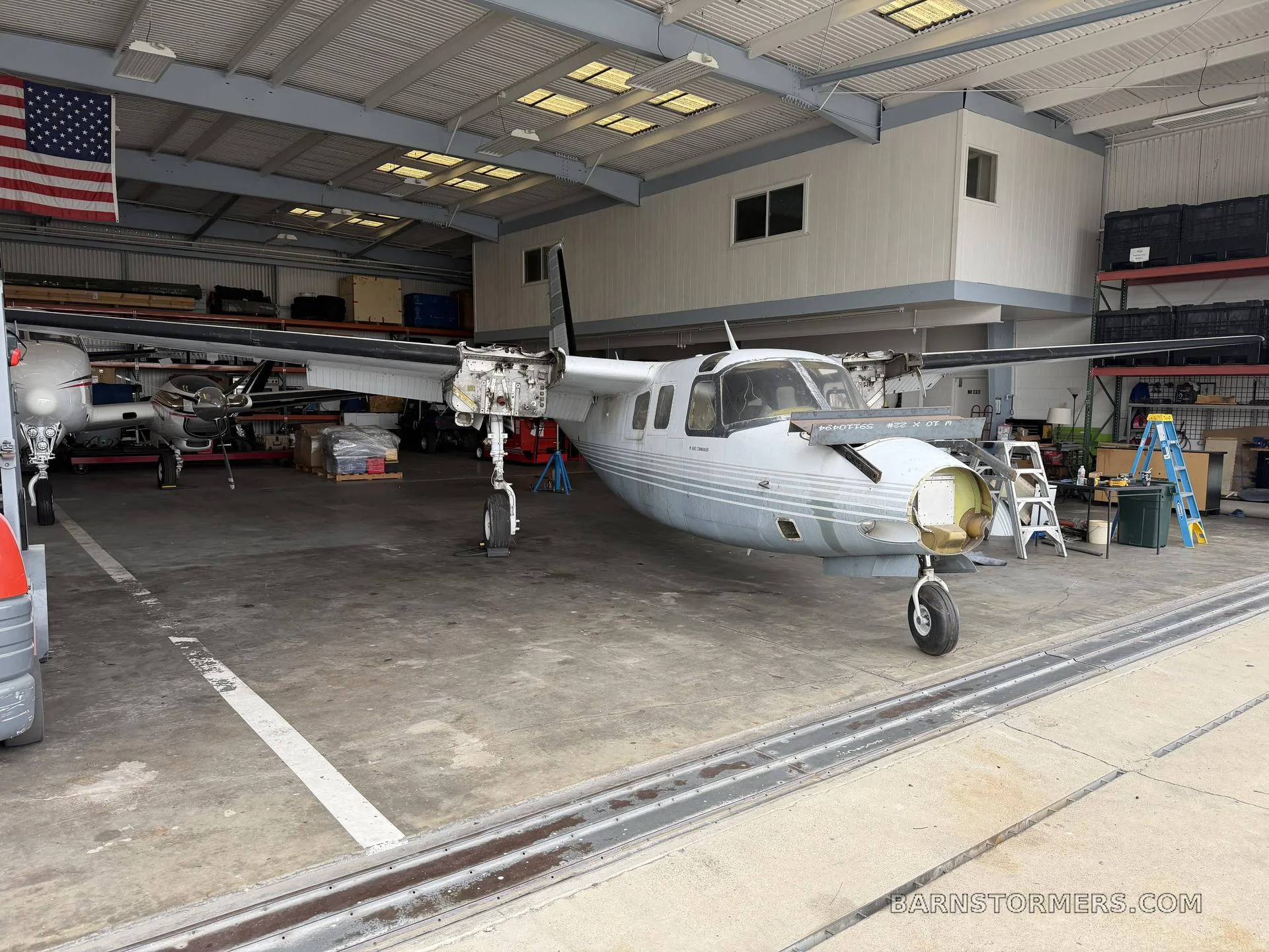 A white twin-engine aircraft with its nose cone and engine cowlings removed, undergoing maintenance in a hangar with an American flag.