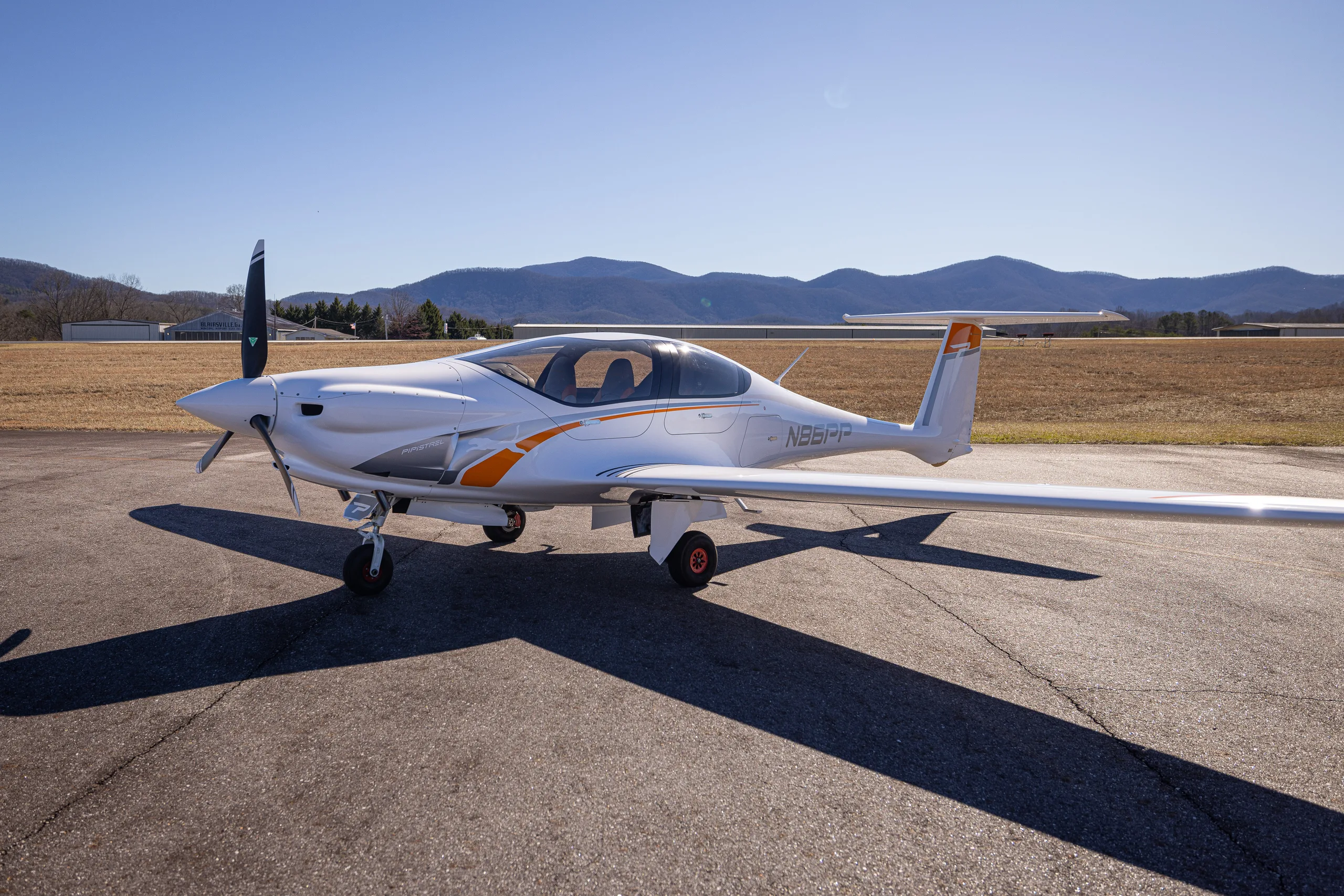 A white Pipistrel aircraft with orange and grey accents is parked on an airfield with mountains in the background.