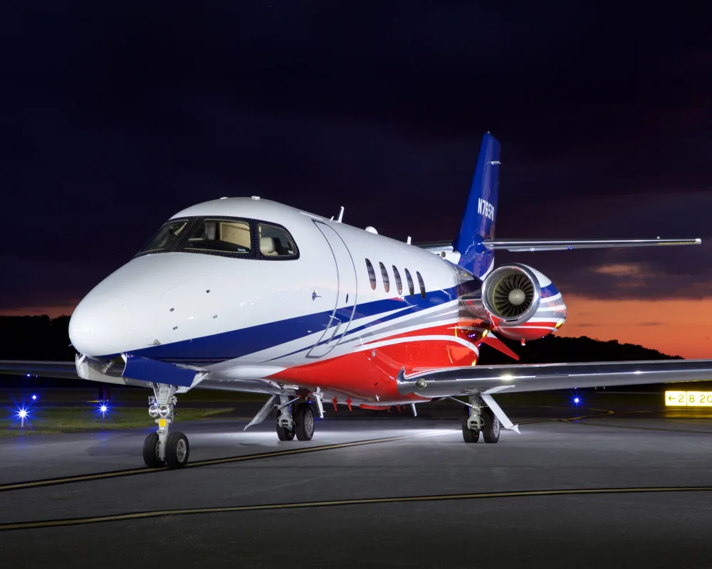 A front-quarter view of a white, red, and blue private jet (N765PG) parked on an airport tarmac at night.
