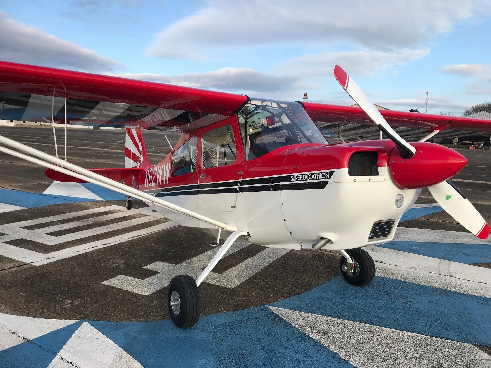 A red and white Super Decathlon aircraft, registration N62WW, parked on a blue and white marked tarmac.