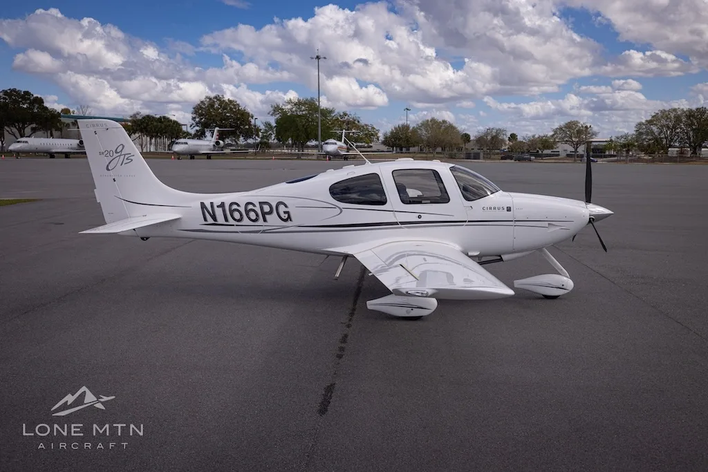 A side view of the white Cirrus SR20 aircraft (N166PG) parked on an airport tarmac with other planes in the background.