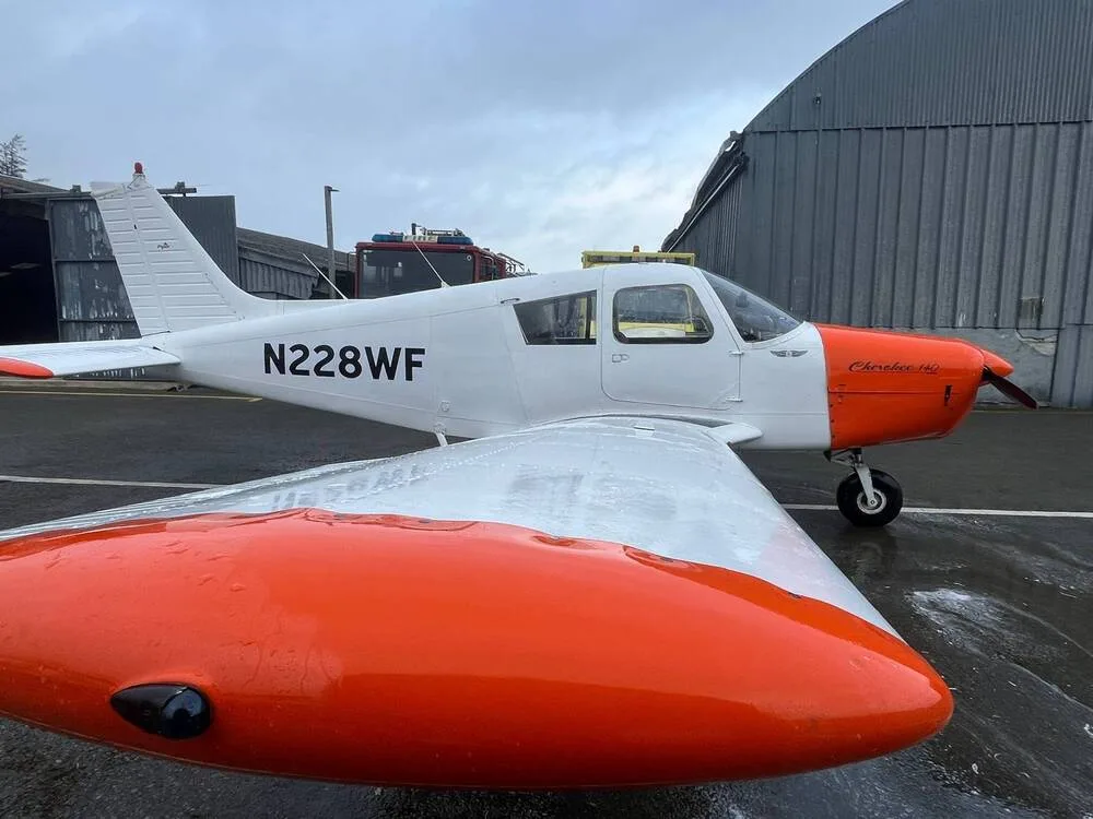 A white and orange Piper Cherokee 140 (N228WF) parked on a wet tarmac next to a hangar, viewed from the front-right.