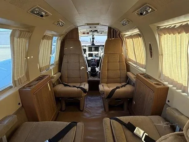 The aircraft cabin interior shows tan seats, wood-grain side panels with cup holders, and window curtains, looking forward towards the cockpit.